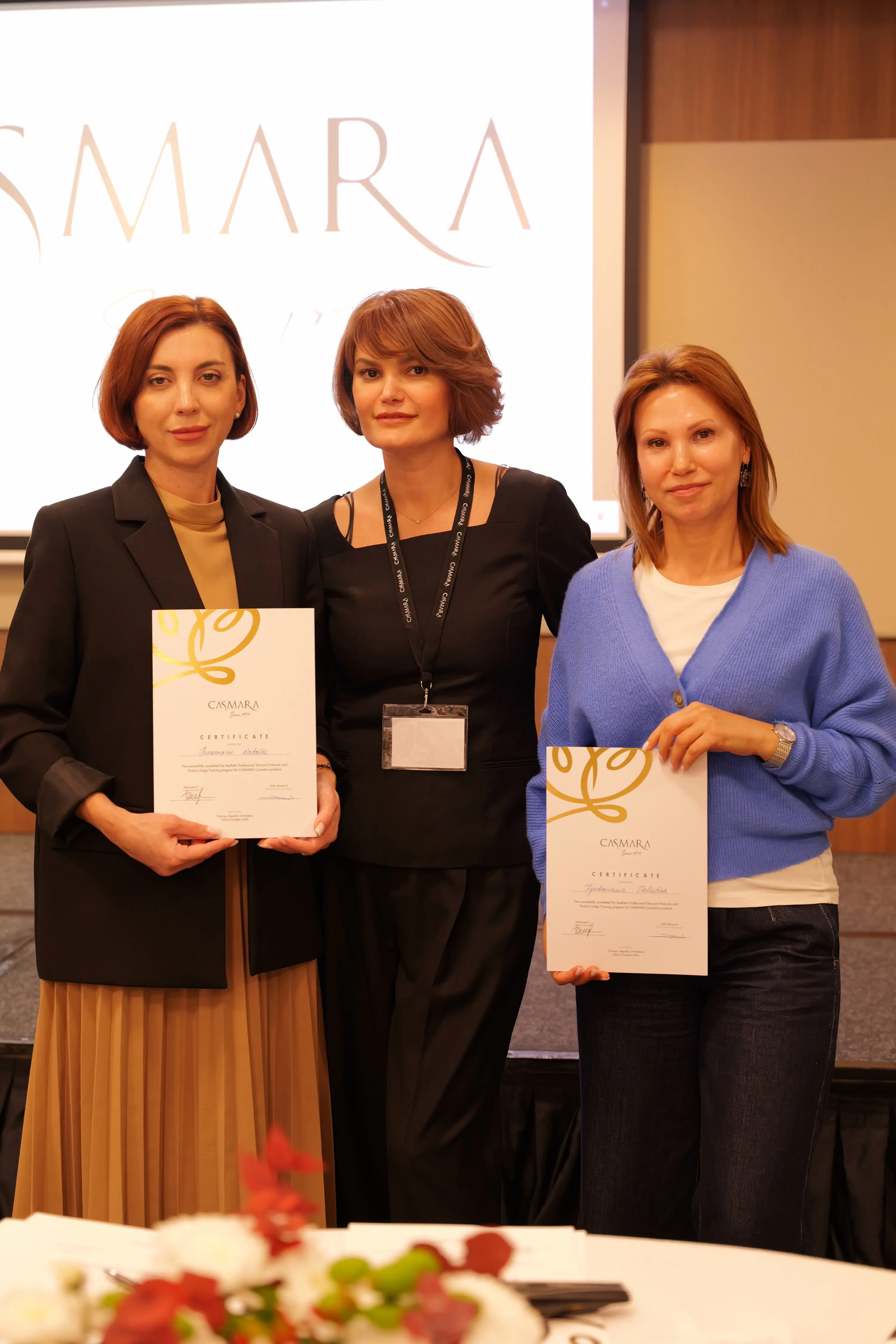 Două participante cu certificatele Casmara, alături de o reprezentantă a brandului; în spate, sigla pe ecran